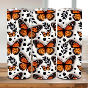 May include: Three white insulated tumblers with a repeating pattern of orange monarch butterflies, black dots, and black leaf sprigs. The butterflies have black and white details. The tumblers are displayed on a wooden surface.