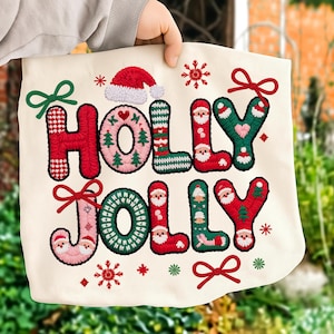 May include: Cream tote bag embroidered with 'HOLLY JOLLY' in red and green. The letters feature Christmas designs like Santa faces, snowflakes, and Christmas trees. A red Santa hat is at the top. Festive and seasonal.