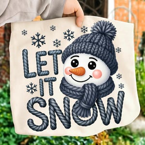 Puede incluir: Bolso tote de color crema con un diseño de muñeco de nieve con gorro y bufanda de punto azul. El muñeco de nieve tiene una nariz de zanahoria, mejillas rosadas y el texto "LET IT SNOW" con copos de nieve.