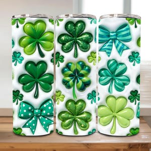 May include: Three stainless steel tumblers with a white background and a repeating pattern of green shamrocks and bows. The shamrocks vary in shades of green, some with glitter. The bows have polka dots and stripes.