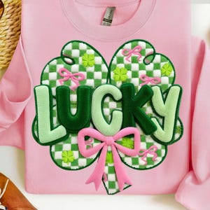 May include: Pink sweatshirt with a large, embroidered shamrock design. The shamrock features a green and white checkered pattern, green and pink bows, and the word "LUCKY" in green and light green. A festive St. Patrick's Day theme.