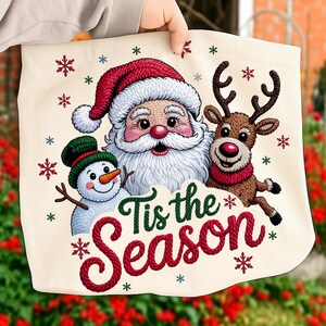 Puede incluir: Un bolso tote de color crema con un diseño navideño festivo. Representa a Papá Noel, un muñeco de nieve y un reno, junto con la frase "Tis the Season" en rojo y verde. Copos de nieve están dispersos alrededor de los personajes.