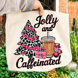 Puede incluir: Bolso tote color crema con un diseño de árbol de Navidad con estampado de leopardo en blanco y negro y lazos rosas. También se representan una taza de café, regalos y adornos. Las palabras "Jolly and Caffeinated" están escritas en negro.