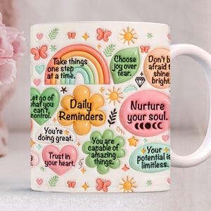 May include: White ceramic mug decorated with colorful inspirational quotes and graphics. The mug features a rainbow, butterflies, suns, and floral elements. Text includes "Daily Reminders" and other positive affirmations.