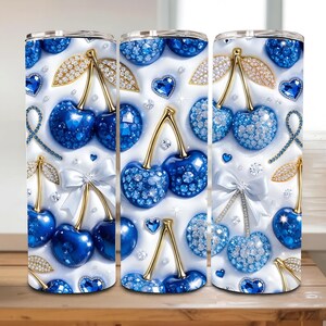 May include: Three stainless steel tumblers featuring a repeating pattern of blue cherries, gold stems, and diamond-like accents on a white background. The design includes white bows and heart-shaped gems. The tumblers are displayed on a wooden surface.