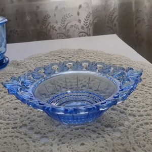 Puede incluir: Un cuenco de vidrio azul con un borde festoneado decorativo. El cuenco tiene forma cuadrada con esquinas redondeadas y un patrón detallado alrededor del borde. El cuenco está sobre un tapete de encaje blanco.