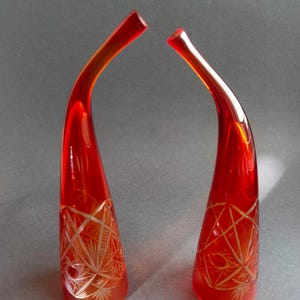 Könnte beinhalten: Zwei hohe, geschwungene rote Glasvasen mit breitem Boden, die sich zu einer schmalen Öffnung verjüngen. Die unteren Bereiche beider Vasen weisen komplizierte, geätzte Stern- und geometrische Muster auf, die mit der glatten, roten Oberfläche kontrastieren.