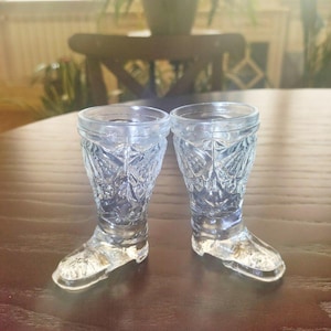 Könnte beinhalten: Zwei klare, geformte Schnapsgläser aus Glas in Form von Cowboy-Stiefeln. Die Gläser haben ein detailliertes, strukturiertes Muster und werden auf einer dunkelbraunen Oberfläche präsentiert. Die Schnapsgläser sind hellblau.