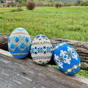 May include: Three beaded Easter eggs in blue, white, and gold sit on a weathered wooden surface. The eggs display detailed patterns, including geometric designs and snowflake motifs, against a backdrop of green fields and a distant mountain.