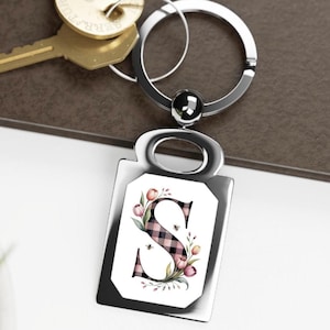 Puede incluir: Un llavero plateado con un colgante rectangular con la letra "S" en un diseño de cuadros rosa y negro, adornado con motivos florales y de abejas. El llavero incluye un llavero circular y un pequeño detalle redondo.