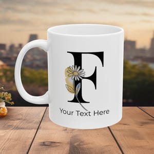 Pode incluir: Caneca de cerâmica branca com a letra preta "F", decorada com uma margarida e uma folha. A caneca tem o texto "Your Text Here". A caneca está sobre uma superfície de madeira com um fundo urbano desfocado.