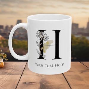 Può includere: Tazza in ceramica bianca con la lettera nera "H" decorata con una margherita e una farfalla. La tazza presenta la scritta "Your Text Here". La tazza è su una superficie di legno con uno sfondo urbano sfocato.