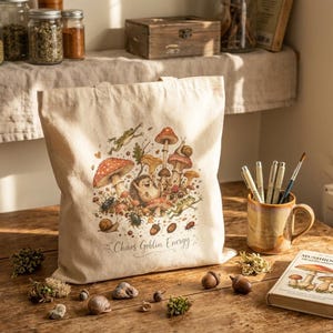 May include: A beige canvas tote bag featuring a colorful illustration of a hedgehog, various mushrooms, frogs, and snails. The text "Chaos Goblin Energy" is printed in a cursive script below the central graphic design on the front of the bag.