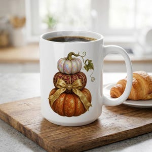 May include: A white ceramic mug featuring a graphic of three stacked pumpkins. The top pumpkin is iridescent, the middle is dark red with gold patterns, and the bottom is orange. A gold ribbon with a heart charm is tied around the middle pumpkin.