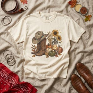 Könnte beinhalten: Ein cremefarbenes Kurzarm-T-Shirt mit einem Grafikdruck aus einem braunen Cowboyhut, braunen Lederstiefeln, Sonnenblumen, Weizenähren, Kürbissen und einer leuchtenden Laterne. Das T-Shirt hat einen Rundhalsausschnitt und ein Etikett am Nacken.