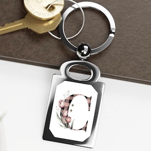 Puede incluir: Un llavero plateado con un colgante rectangular. El colgante presenta la letra "C" en un patrón de cuadros rosa y negro, rodeada de tulipanes rosas y hojas verdes. Un llavero plateado y algunas llaves son visibles.