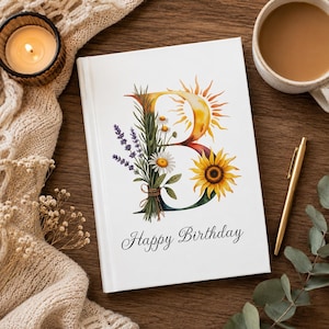 May include: A white journal with a floral design featuring a yellow sun, sunflower, and the letter "B". The words "Happy Birthday" are written in script below the design. A lit candle, a cup of coffee, and a gold pen are also visible.