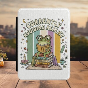 May include: A white e-reader case featuring a green frog wearing glasses and reading a book titled Dragon's Tale. The illustration includes a stack of books labeled Myths, Magic, Legends, Tales, and Fantasy, with the text Currently Avoiding Reality.