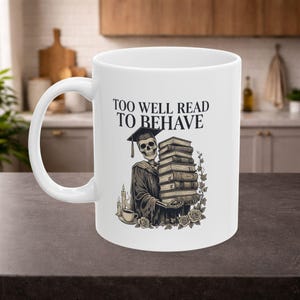 Puede incluir: Taza de cerámica blanca con un esqueleto con toga de graduación sosteniendo libros. La taza tiene el texto "TOO WELL READ TO BEHAVE". El diseño incluye rosas y velas. Ideal para bebidas calientes.