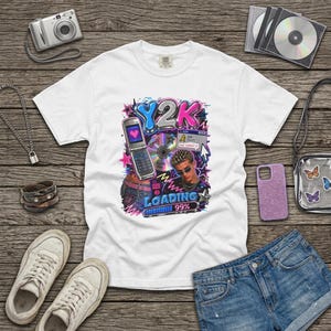 May include: A white short-sleeve t-shirt featuring a Y2K-themed graphic print. The design includes a flip phone, a compact disc, a person with spiky hair wearing sunglasses, and a loading bar at 99 percent with the text Y2K and AIM.
