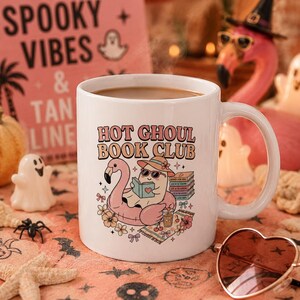 Op de afbeelding: Witte keramische mok met de tekst "HOT GHOUL BOOK CLUB" en een cartoonillustratie van een spook dat een boek leest op een flamingo-drijver. De mok is omgeven door Halloween-thema decoraties.