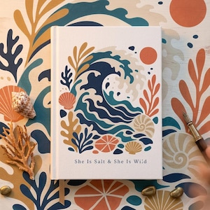 May include: A white hardcover journal featuring a central illustration of a large blue ocean wave surrounded by coral, seashells, and a sun in shades of orange, yellow, and navy blue. The text "She Is Salt & She Is Wild" is printed at the bottom.