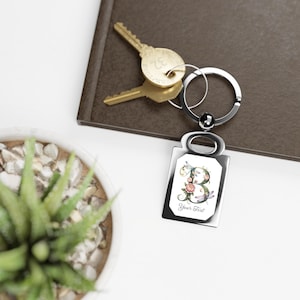 May include: A silver rectangular keychain with a floral letter "B" design. The letter is adorned with pink roses, green leaves, and dragonflies. The keychain also has the text "Your Text" below the letter.
