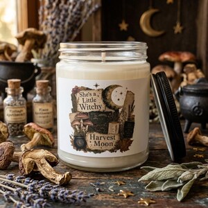 Pode incluir: Um frasco de vela de vidro transparente com cera branca e tampa preta. O rótulo apresenta uma lua crescente, cogumelos e um caldeirão com o texto "She's a Little Witchy" e "Harvest Moon" impresso numa fonte decorativa de estilo vintage.