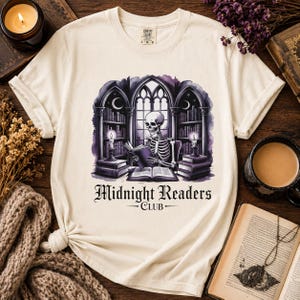 Könnte beinhalten: Cremefarbenes T-Shirt mit einer Grafik eines Skeletts, das in einer gotischen Bibliothek liest. Das Design umfasst Bücherregale, Kerzen und den Text "Midnight Readers Club". Das T-Shirt ist aus weichem Material. Das Design ist in Violetttönen gehalten.