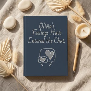 May include: A dark blue hardcover journal featuring the white text "Olivia's Feelings Have Entered the Chat." and a graphic of two speech bubbles containing heart shapes. The journal is positioned on a neutral fabric surface with decorative elements.