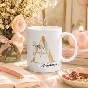May include: White ceramic mug featuring a watercolor letter "A" in pastel colors, adorned with a bow. The name "Annalisa" is printed below the letter. The mug has a curved handle and is set on a table.