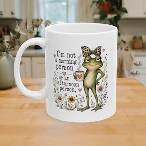 Puede incluir: Una taza de cerámica blanca con una rana verde que lleva un lazo con estampado de leopardo y sostiene una pequeña taza de café. El texto dice, I'm not a morning person or an afternoon person, rodeado de corazones rosas y flores silvestres.
