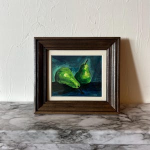 May include: A framed painting of two green pears on a dark blue background. The pears are illuminated, with highlights and shadows. The frame is dark brown, and the painting is displayed on a marble surface.