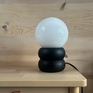 May include: A table lamp featuring a large, spherical white glass shade resting on a base composed of two stacked, matte black circular rings. A black power cord extends from the base, which is positioned on a light-colored wooden surface.