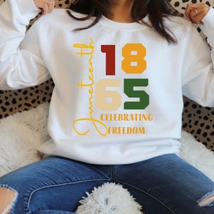 May include: White sweatshirt with the words "Juneteenth" in yellow script. The numbers "1865" are in red, gold, and green. The words "Celebrating Freedom" are below the numbers.