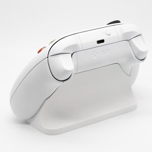 May include: A white Xbox controller resting on a matching white charging stand. The controller has black buttons and triggers, with the Xbox logo visible. The stand is designed to cradle the controller.