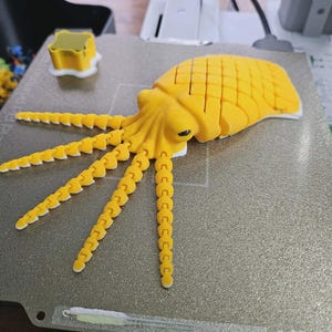 May include: A yellow 3D-printed octopus model with segmented body and tentacles, resting on a textured silver surface. The model has a detailed design, including a textured head and eye. A small yellow square is also visible.