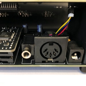 K&ouml;nnte beinhalten: Nahaufnahme des Inneren eines elektronischen Ger&auml;ts. Das Bild zeigt Leiterplatten, einen runden MIDI-Anschluss, einen Stromeingang und eine Audiobuchse. Gelbe, rote und blaue Dr&auml;hte sind sichtbar.