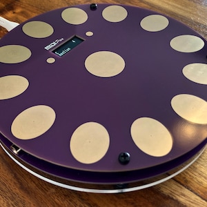 Könnte beinhalten: Ein kreisförmiger violetter MIDI-Controller mit zwölf goldfarbenen, kreisförmigen Touchpads, die in einem Ring angeordnet sind, sowie einem zentralen Pad. Das Gerät verfügt über ein kleines Digitaldisplay mit dem Text MIDI Pan und Aeolian.