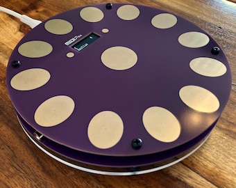 Puede incluir: Un controlador MIDI circular de color morado con doce almohadillas táctiles circulares doradas dispuestas en anillo y una central. El dispositivo cuenta con una pequeña pantalla digital que muestra el texto MIDI Pan y Aeolian, con un cable blanco.