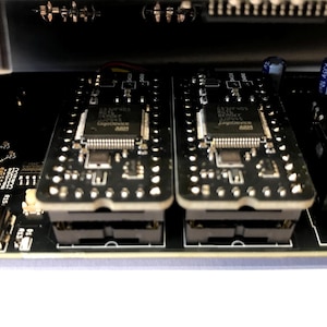 K&ouml;nnte beinhalten: Nahaufnahme von elektronischen Bauteilen, darunter zwei schwarze Leiterplatten mit integrierten Schaltkreisen und Stiften. Die Platinen sind in einem schwarzen Geh&auml;use montiert, mit einem USB-Anschluss und Kondensatoren.