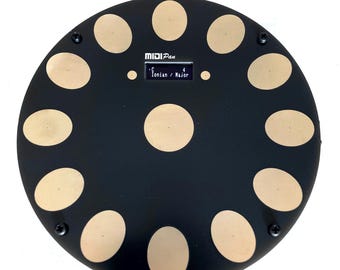 Peut inclure: Un contrôleur MIDI noir circulaire avec douze pavés tactiles beiges disposés en cercle et un pavé central. L'appareil comporte un petit écran numérique affichant le texte MIDIPan et C 4 Ionian / Major, avec un câble blanc sur le dessus.