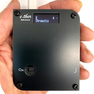 Könnte beinhalten: Ein schwarzes elektronisches Gerät mit dem Text "e-ther Advance" und einem kleinen Bildschirm mit der Anzeige "E Chromatic 4." Das Gerät hat eine Taste und wird in einer Hand gehalten. Ein weißes Kabel ist oben eingesteckt.