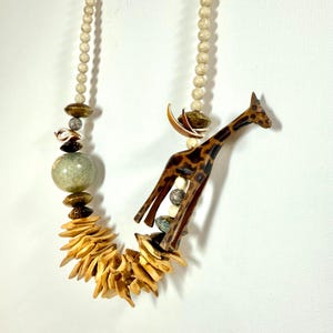May include: A unique necklace featuring a carved wooden giraffe pendant with brown spots, accented by a variety of natural beads, shells, and wooden elements. The necklace includes a strand of small, round, off-white beads and a cluster of light brown, petal-shaped pieces.