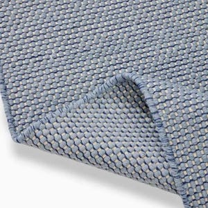 Puede incluir: Primer plano de una alfombra texturizada azul y blanca. La alfombra tiene un patrón tejido con un fondo azul claro y detalles blancos. El borde de la alfombra está doblado, revelando la parte inferior.