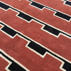 May include: Close-up of a rug with a geometric pattern. The rug features a repeating design of black and white shapes against a reddish-orange background. The texture appears soft and plush, suggesting a comfortable floor covering.