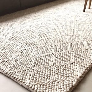 以下が含まれることがあります： 幾何学模様のベージュ色のテクスチャード加工されたラグ。 ラグは、織り込まれた要素と盛り上がった要素を組み合わせており、触感のある表面を作り出しています。 ラグは長方形で、天然繊維で作られているようです。