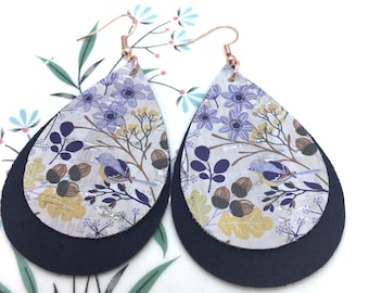 May include: A pair of teardrop-shaped earrings with a layered design. The top layer is a white background with a floral pattern in shades of purple, blue, and yellow. The bottom layer is a solid navy blue. The earrings have a rose gold hook.