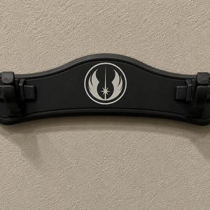 K&ouml;nnte beinhalten: Eine schwarze Wandhalterung f&uuml;r ein Lichtschwert mit geschwungener Form. Sie hat ein wei&szlig;es Emblem in der Mitte und zwei Clips zur Befestigung von Lichtschwertern. Die Halterung ist an einer strukturierten, hellfarbigen Wand befestigt.