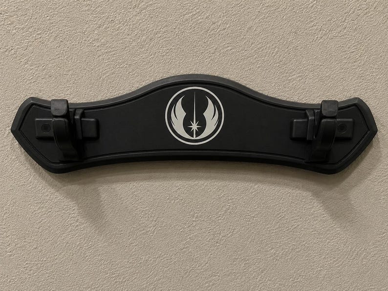 K&ouml;nnte beinhalten: Eine schwarze Wandhalterung f&uuml;r ein Lichtschwert mit geschwungener Form. Sie hat ein wei&szlig;es Emblem in der Mitte und zwei Clips zur Befestigung von Lichtschwertern. Die Halterung ist an einer strukturierten, hellfarbigen Wand befestigt.